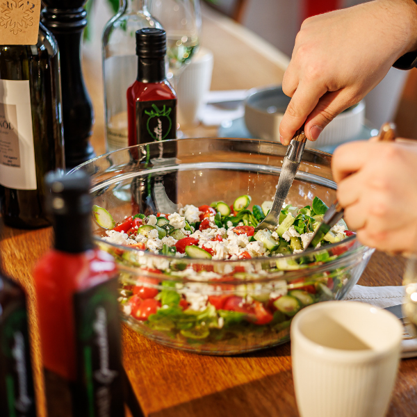 Ein frischer Salat aus Tomaten, Gurken und Feta wird in einer Glasschüssel zubereitet. Eine Hand hält zwei Utensilien, um die Zutaten zu vermengen. Im Hintergrund sind Flaschen mit Öl und Sauce sichtbar, die eine einladende Essenssituation schaffen.