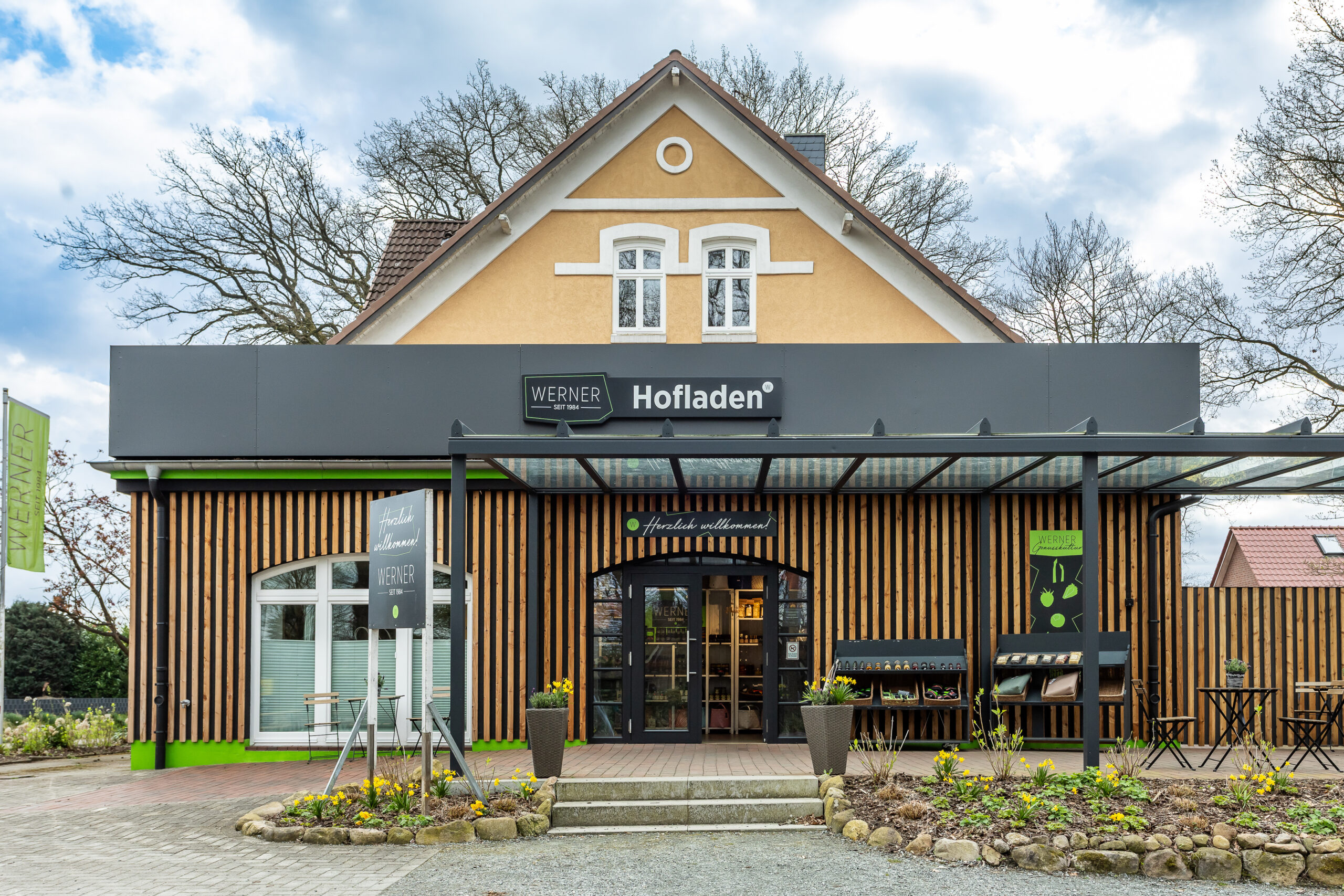 Hofladen von Werner, ein einladendes Gesch&auml;ft mit modernem Design, zeigt eine Holzfassade und gro&szlig;e Fenster. Vor dem Eingang stehen bl&uuml;hende Pflanzen und Tische mit St&uuml;hlen, umgeben von einem gepflegten Garten. Das Schild &uuml;ber dem Eingang weist auf die Produkte des Hofladens hin und vermittelt eine einladende Atmosph&auml;re.